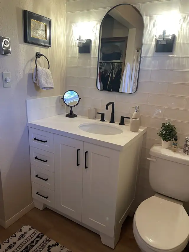 Updated bathroom with white shaker vanity, black faucet and pulls, and a white tile wall behind the arched mirror